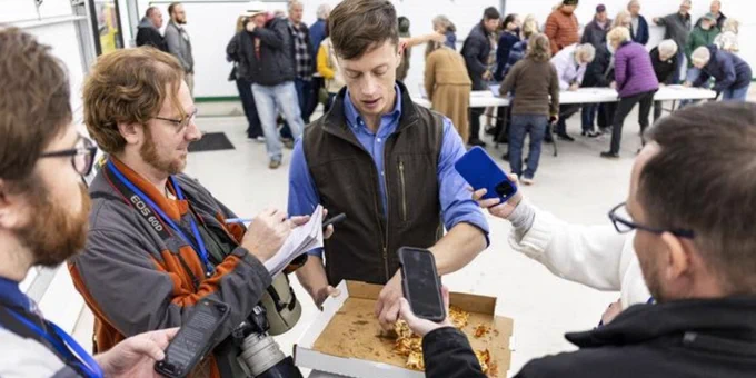 A photo of Rob Sand speaking to the press and eating Mabe's pizza during his 2025 Town Hall Tour stop in Decorah.