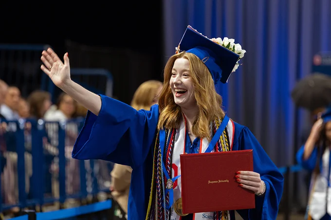 Skyler waves to the crowd after receiving her diploma. She is wearing her blue cap and gown along with many cords and shashes.