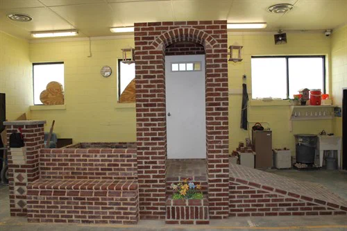 Indoor room with a central brick structure featuring a white door and flower bed at its base, surrounded by yellow walls and windows.