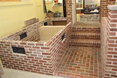 Indoor brick structure with a sunken area and steps leading down, surrounded by yellow walls and workshop tools in the background.