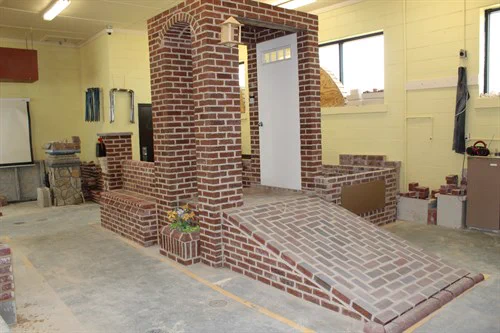 Indoor brick structure with a ramp leading to a white door, surrounded by brick walls and a small flower planter on the side.