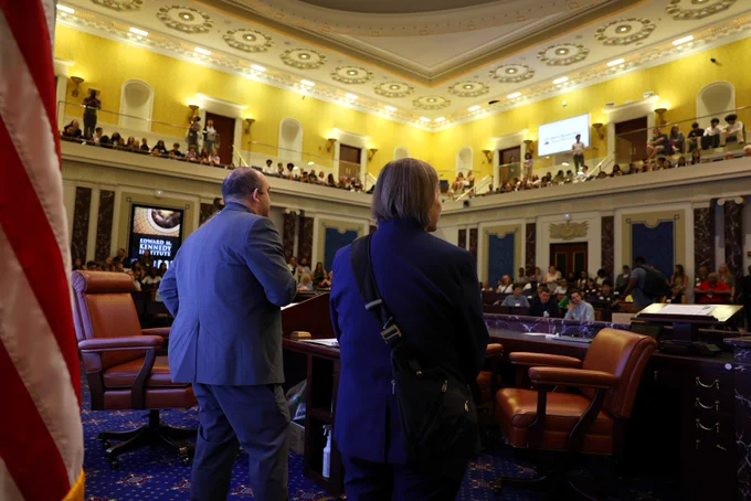 Massachusetts Education Secretary Steve Zrike addresses students gathered inside the Edward M. Kennedy Institute’s Senate Chamber during DESE’s Civics Project Showcase. Viewed from behind the podium area, Zrike stands before a packed audience of students seated throughout the chamber and along the upper gallery, beneath the room’s ornate gold ceiling and historic architecture.