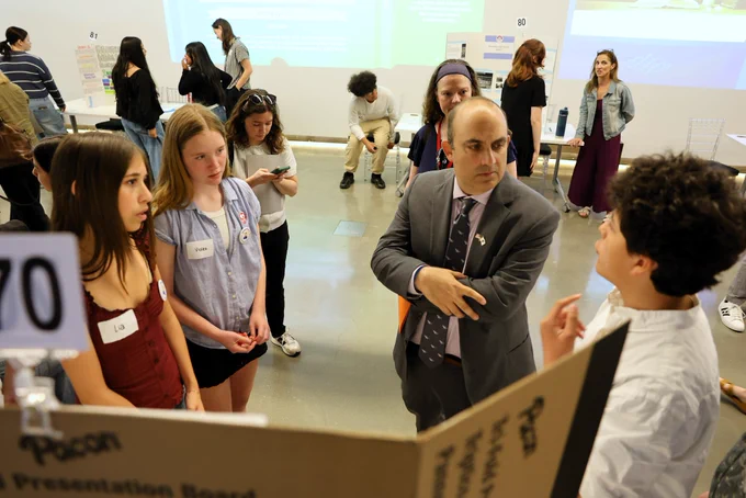 Massachusetts Education Secretary Steve Zrike listens as students present their civics project during DESE’s Civics Project Showcase at the Edward M. Kennedy Institute. Standing with a group of students around a presentation board, Zrike engages in conversation while students explain their research and findings in a bright exhibition space filled with project displays and participants.