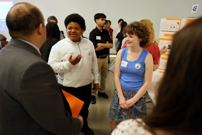 Massachusetts Education Secretary Steve Zrike listens as students present their civics project during DESE’s Civics Project Showcase at the Edward M. Kennedy Institute. Standing with a group of students around a presentation board, Zrike engages in conversation while students explain their research and findings in a bright exhibition space filled with project displays and participants.