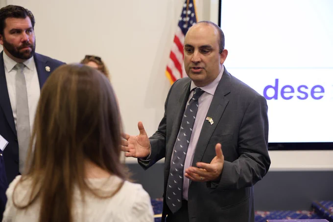 Secretary Zrike in a suit and tie speaks with a student during the Civics Projects Showcase. An American flag and the DESE logo can be seen behind them.