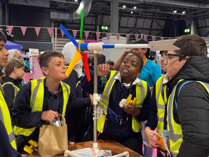 A group of young students wearing reflective vests are gathered around a colourful wind turbine model. They are actively discussing and pointing at the turbine, which is made of a sturdy base and vibrant blades.