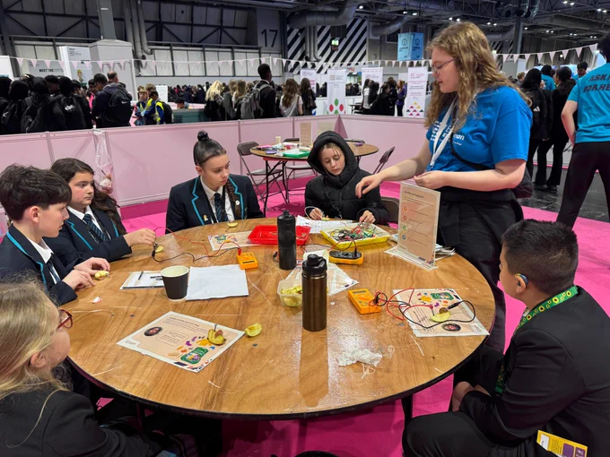 A group of young people sit around a round table engaging in a science activity, using fruits and wires to conduct an experiment. One person stands explaining, wearing a blue shirt. The setting appears to be an educational event or workshop in a large hall with many attendees and booths visible in the background.