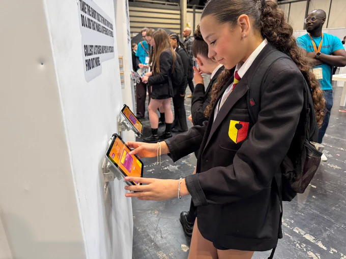 A person in a school uniform interacts with a station displaying multiple iPads mounted horizontally. Each screen shows a bright, colourful interface. Several other individuals, also in school attire, engage with their own phones at the same station. A staff member stands nearby, observing.