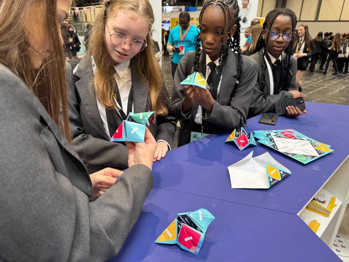 A group of students in school uniforms engages with colorful paper fortune tellers at an event. They appear focused and attentive. The background shows a bustling event space with other attendees.