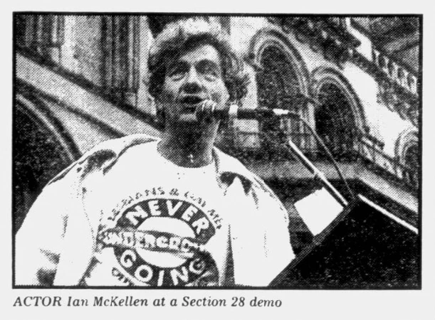 Black and white photograph of Ian McKellan speaking at a podium at a Section 28 demo. He wears a T-shirt with a slogan, and a jacket.
