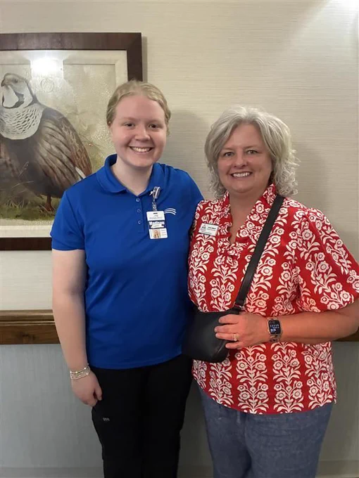 Two women stand smiling together indoors. On the left is Surry-Yadkin Works graduate Madison Moorefield, wearing a blue RidgeCrest polo shirt with an employee badge. On the right is Candy from Surry-Yadkin Works, wearing a red-and-white patterned blouse. The photo was taken during a visit celebrating Madison's one-year work anniversary at RidgeCrest. A framed wildlife print hangs on the wall behind them.