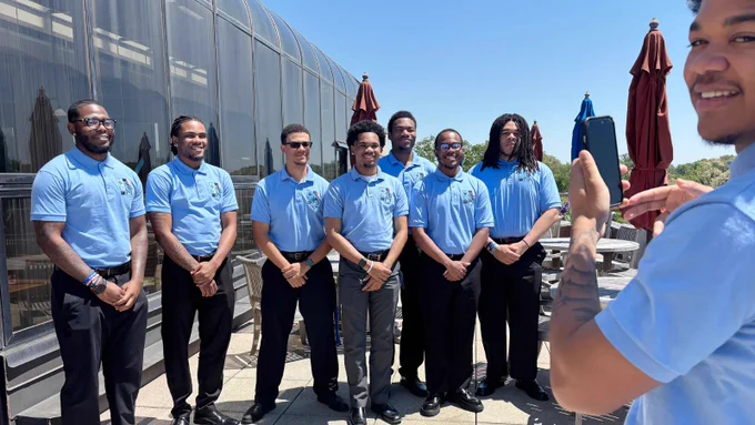 A group of young black men in matching sky blue polos stand together with their hands clasped posing for a picture.