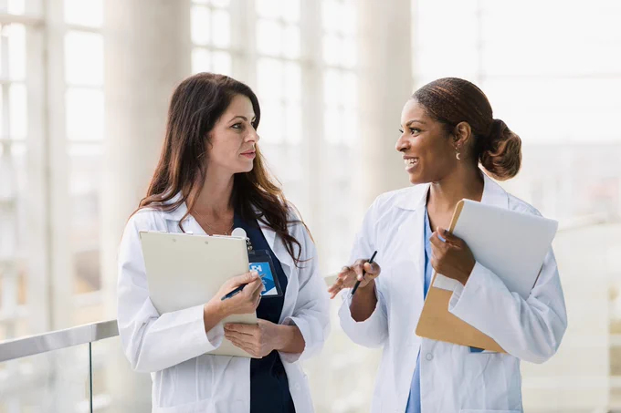 Two medical professionals in white coats stand indoors, engaged in conversation. They appear to be discussing something important in a well-lit building with large windows.