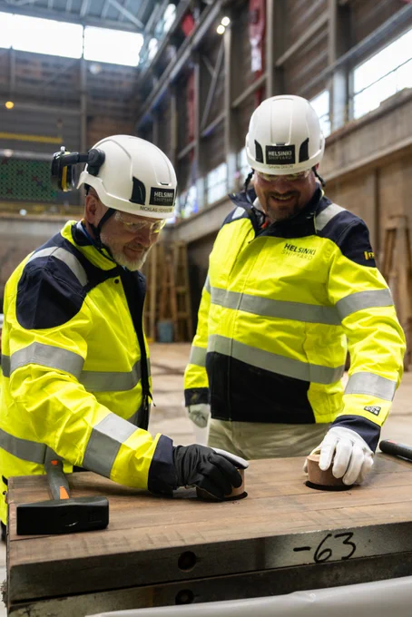 Two workers wearing white hard hats and high-visibility jackets inspect a metal piece in an industrial workshop.