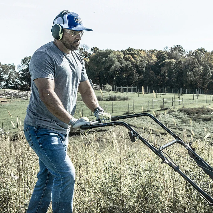 Man wearing ear protection and gloves brush cutting tall grass in a rural field on a sunny day.