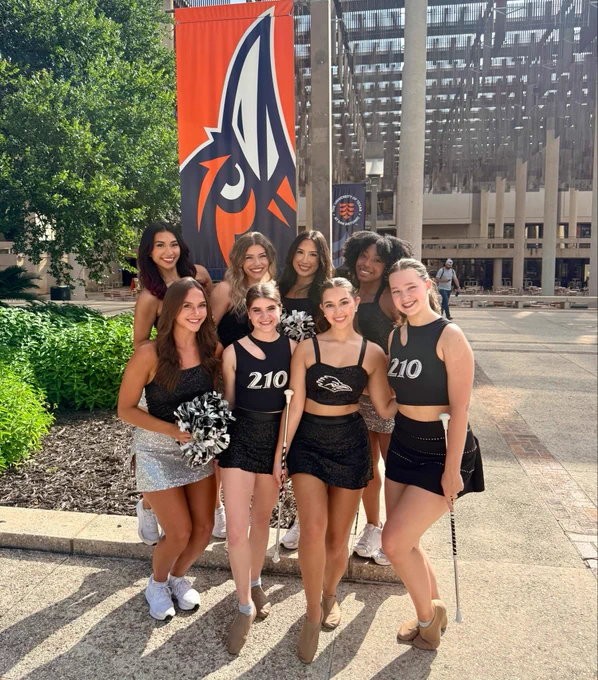 Cheerleaders and twirlers from UTSA stand in front of a Rowdy head logo flag.
