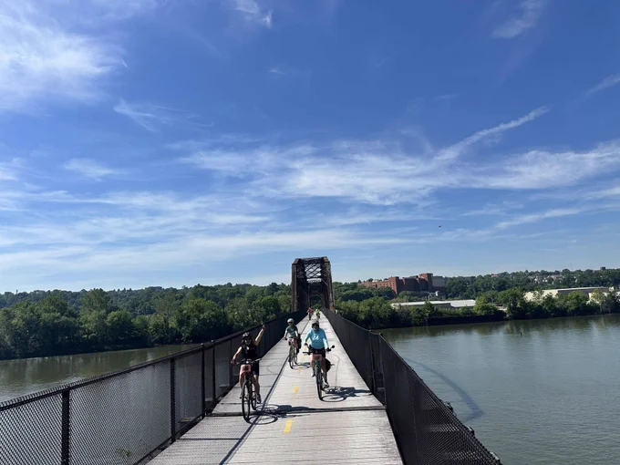 Cyclists riding on a narrow bridge over a calm river under a clear blue sky with trees and buildings in the background.