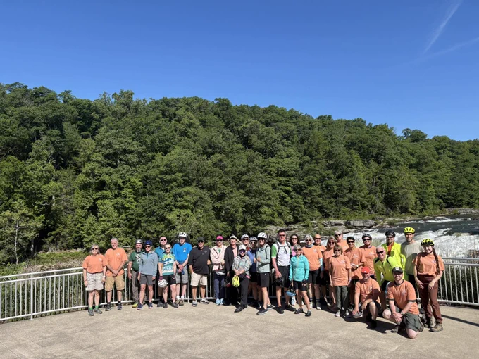 Group of cyclists posing on a riverside platform with lush green forest and flowing water in the background under clear blue sky.