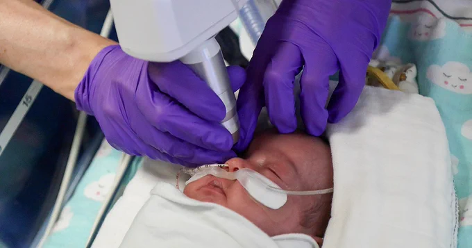 A Duke healthcare worker wearing purple gloves is using a medical scanner to provide care to a sleeping newborn wrapped in a blanket. The newborn is on a bed with a cute cloud-patterned cover and has medical tape on their face with a tube.
