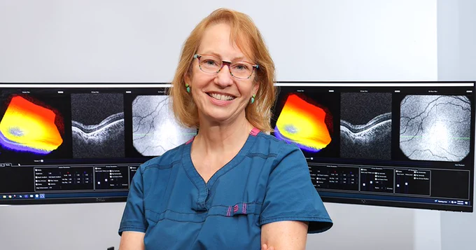 Duke physician Cynthia Toth is in scrubs smiling in front of multiple computer monitors displaying detailed eye scans.