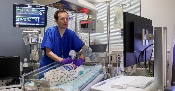 A Duke healthcare professional in a blue uniform attending to a newborn in an incubator surrounded by medical equipment in a hospital room.