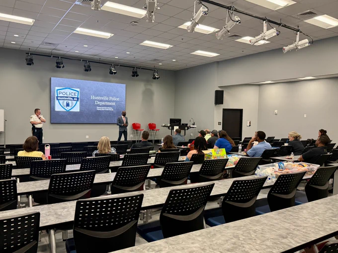 A classroom-style training room contains multiple rows of tables and black perforated chairs facing a presentation area. At the front of the room, a large screen displays the Huntsville Police Department logo and text. Two individuals stand near the screen, one in uniform and one in business attire, addressing the audience. Several attendees sit scattered throughout the room, some with notebooks, snacks, or drinks on the tables. The room is well lit with ceiling-mounted lights and presentation equipment overhead.