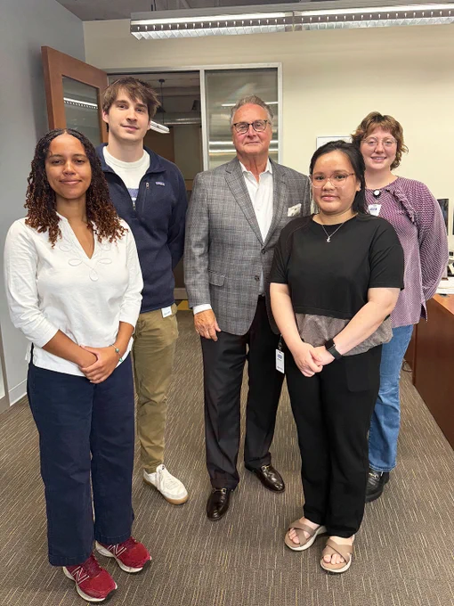 Five professionals standing together in an office setting, dressed in business casual and formal attire, posing for a group photo.