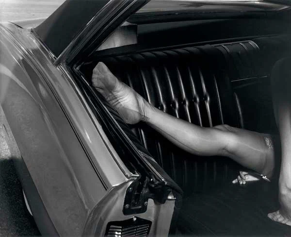 Foto in bianco e nero dall'esterno del finestrino posteriore aperto di un'auto d'epoca con sedili in pelle nera lucida a costine. All'interno, una gamba femminile sollevata poggia sul sedile. La donna indossa calze velate in nylon con la classica riga posteriore scura ben visibile e un reggicalze. L'inquadratura taglia il corpo concentrandosi sulla gamba, sul piede e sulla carrozzeria metallica e riflettente dell'auto.