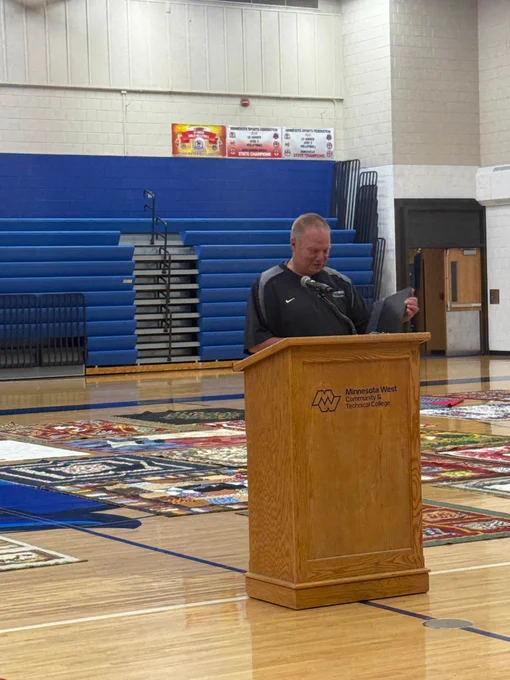 Person speaking at a wooden podium labeled “Minnesota West Community & Technical College” on a gym floor, surrounded by quilts, with blue bleachers and sports banners in the background.