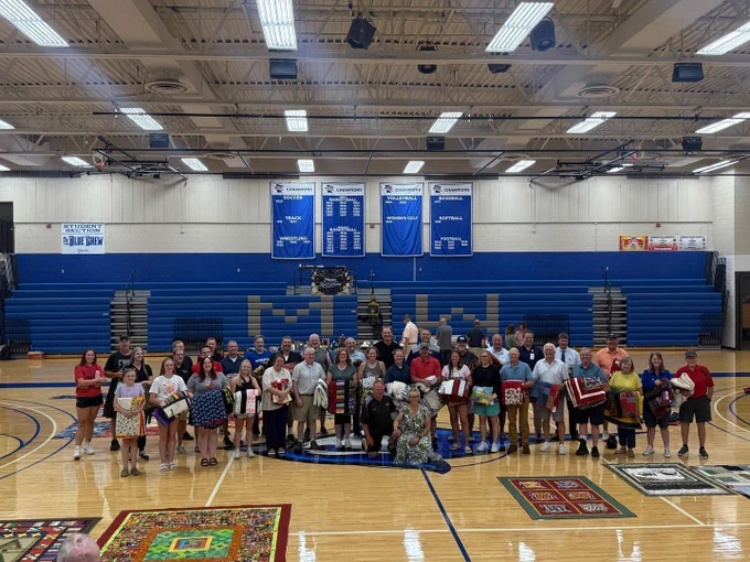 Large group photo in a gymnasium with blue bleachers and championship banners; dozens of people stand on the basketball court holding colorful handmade quilts, while additional quilts are spread out on the floor in front.