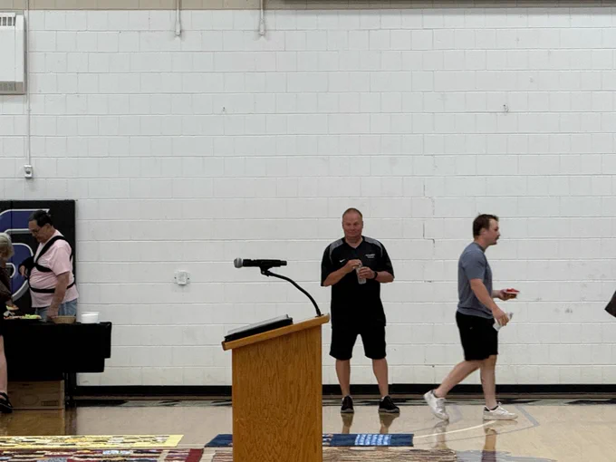 People gather at a table with refreshments inside a gymnasium while a wooden podium and microphone stand at center court.