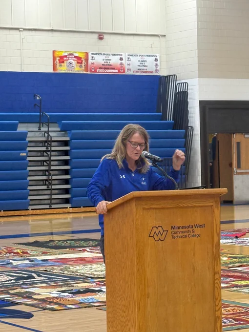 A speaker stands at a wooden podium with a microphone in a gymnasium, with folded bleachers and sports banners in the background.