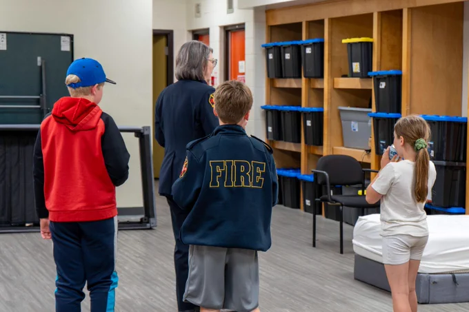 Children and a firefighter standing in a room with storage bins and chairs.