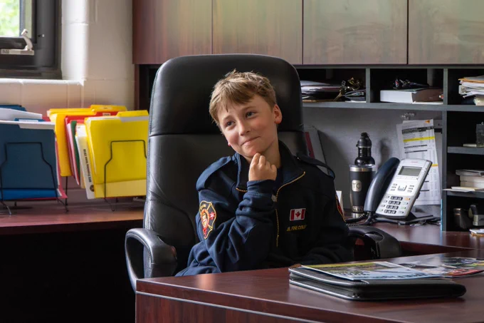 Child in a navy blue uniform with a Canadian flag patch sits thoughtfully in a black office chair behind a wooden desk.