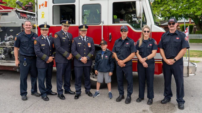 Group of firefighters and a young boy standing in front of a red and white fire truck on a street.