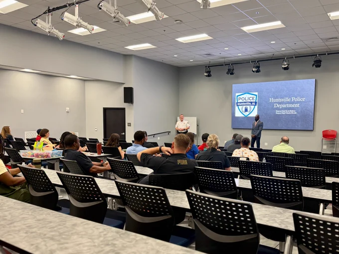 A classroom-style training room contains multiple rows of tables and black perforated chairs facing a presentation area. At the front of the room, a large screen displays the Huntsville Police Department logo and text. Two individuals stand near the screen, one in uniform and one in business attire, addressing the audience. Several attendees sit scattered throughout the room, some with notebooks, snacks, or drinks on the tables. The room is well lit with ceiling-mounted lights and presentation equipment overhead.