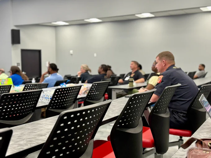 A classroom-style setting shows several individuals seated at long tables with black perforated chairs, facing toward the front of the room. Snacks, drinks, and personal items are scattered on the tables. One individual in a dark uniform sits in the foreground. The room has gray walls, a closed door, and ceiling lights providing even illumination.