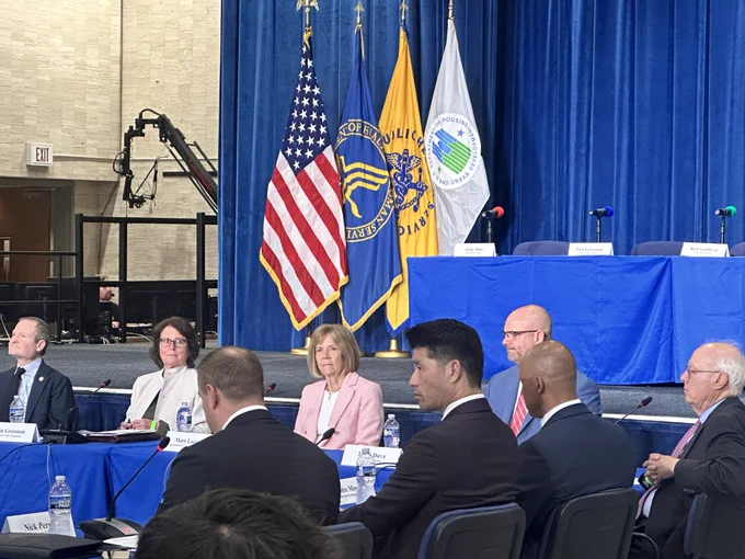 Participants sit around a conference table during a meeting to discuss the Federal Elder Justice Action Plan. Mary Lazare, ACL’s Principal Deputy Administrator, is seated among attendees, with U.S., HHS, and HUD flags displayed on the stage behind them.