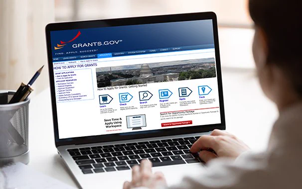 A person is using a laptop displaying the Grants.gov website. The screen shows navigation options and links related to applying for grants.