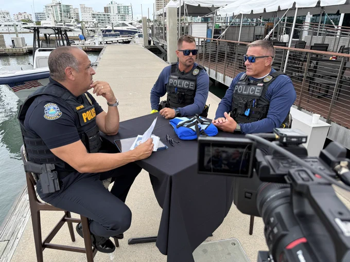 Three police officers in tactical gear sit at a small table on a marina dock. They are engaged in discussion, with a camera filming the scene.