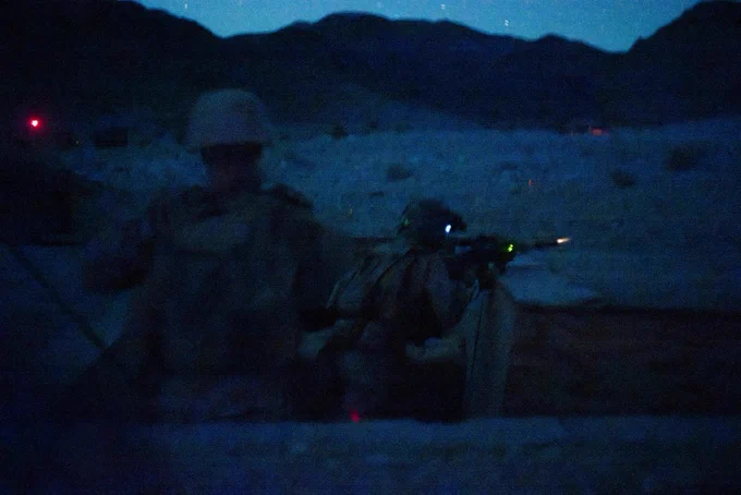 A Marine engages targets during a night live-fire training event while another Marine observes from a nearby fighting position.