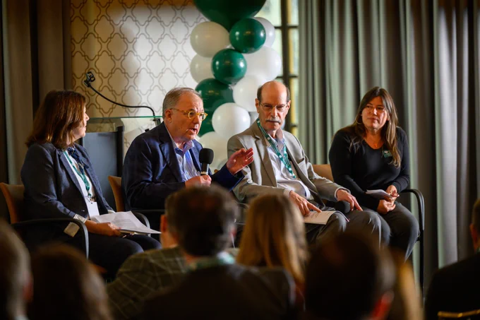 A panel of four people seated on chairs, engaging in a discussion. One person holds a microphone while speaking. Behind them, green and white balloons are arranged. An audience is visible in the foreground.
