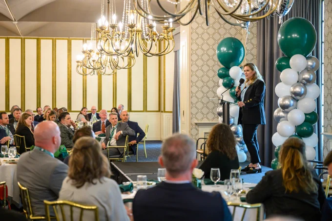 A person is speaking at a podium with an audience seated at tables in a formal event setting. Green and white balloon decorations are visible, and chandeliers hang from the ceiling.