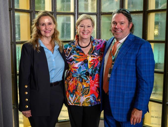 Three individuals stand together in front of a window. The person in the center is wearing a vibrant, floral-patterned jacket. The person on the left is wearing a dark blazer, and the person on the right is in a blue checkered suit. They are all smiling at the camera.