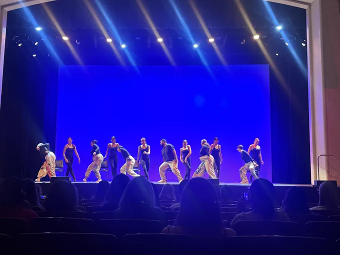 Students perform a choreographed dance on stage under blue lighting, with audience members watching from the theater seats.