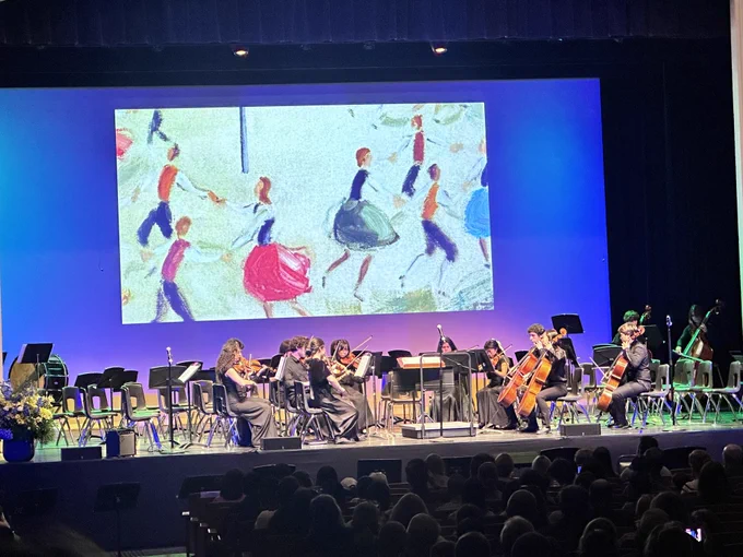 Student orchestra performs on stage before a large audience, with colorful artwork projected on a screen behind the musicians.