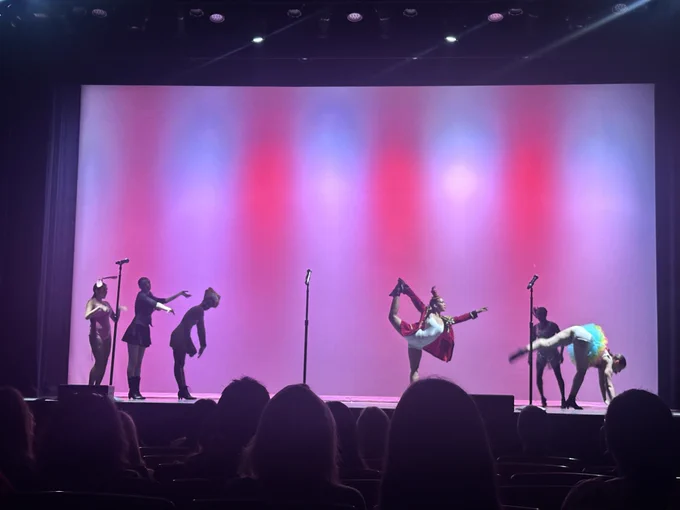 Performers in colorful costumes strike dramatic poses during a stage production illuminated by pink and purple lighting.