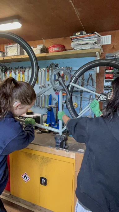 Two people in green gloves working on a bicycle in a workshop, with tools on the wall behind them.
