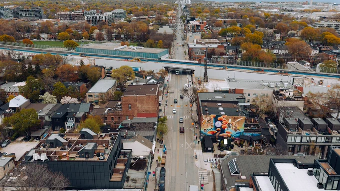 Aerial view of a city street in a residential area with trees showing autumn colors. The street is lined with buildings and leads towards a distant urban skyline. A vibrant mural is visible on one of the buildings. 