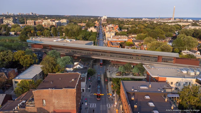 Aerial view of a neighborhood showing a train station and tracks running through the middle, surrounded by buildings and trees. In the distance, there is a tall monument on the right side.